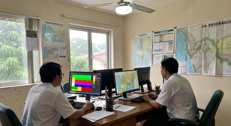 Two staff members at desktop computers showing colorful map layers in a small district office, paper maps pinned on the wall, a ceiling fan, rainy season light through the window.